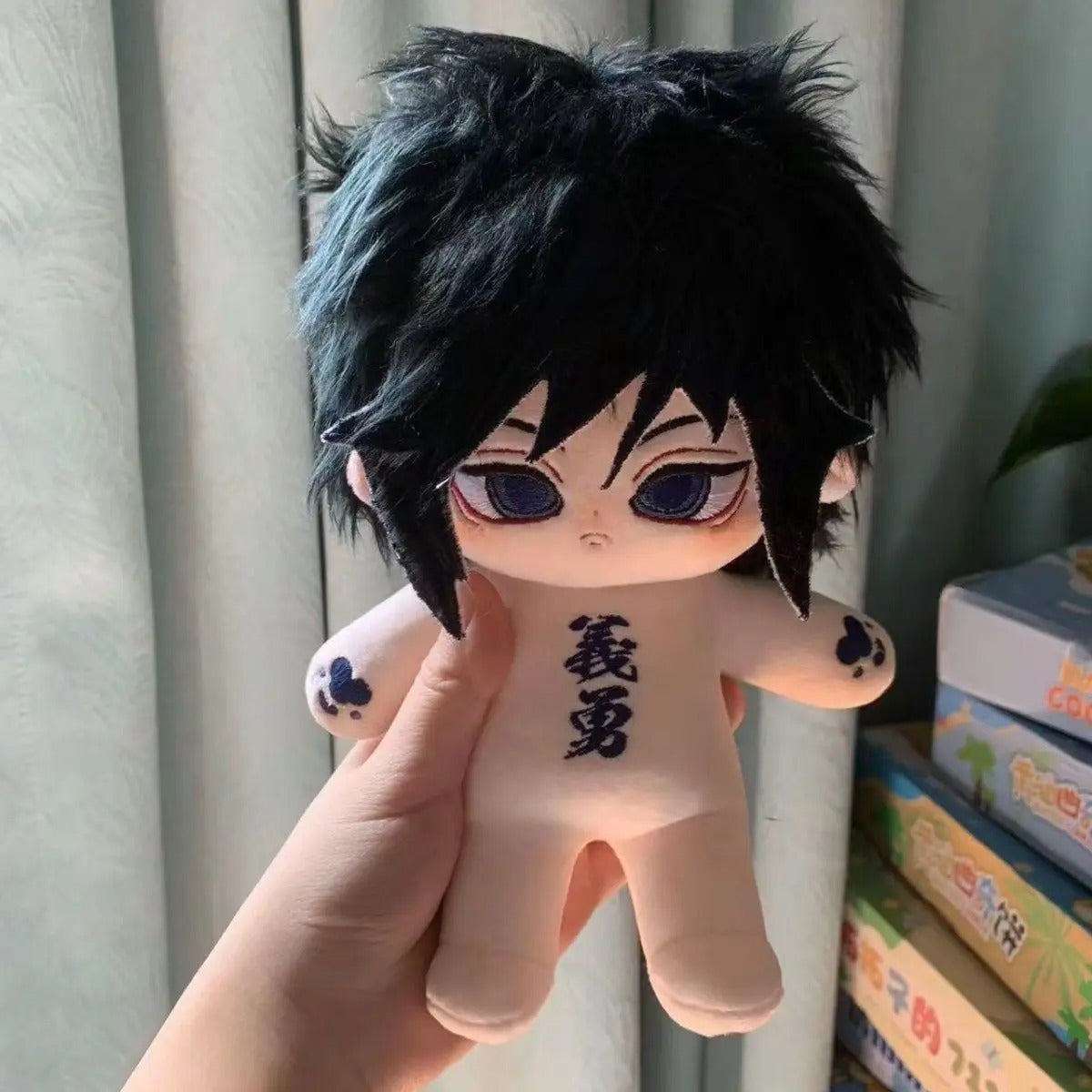 Plush toy with black hair and red eyes held by a hand, with books in the background. Demon Slayer: Kimetsu no Yaiba Giyu Tomioka Premium 20cm Super Soft Plushie Doll – Embroidered Cat Paw Edition