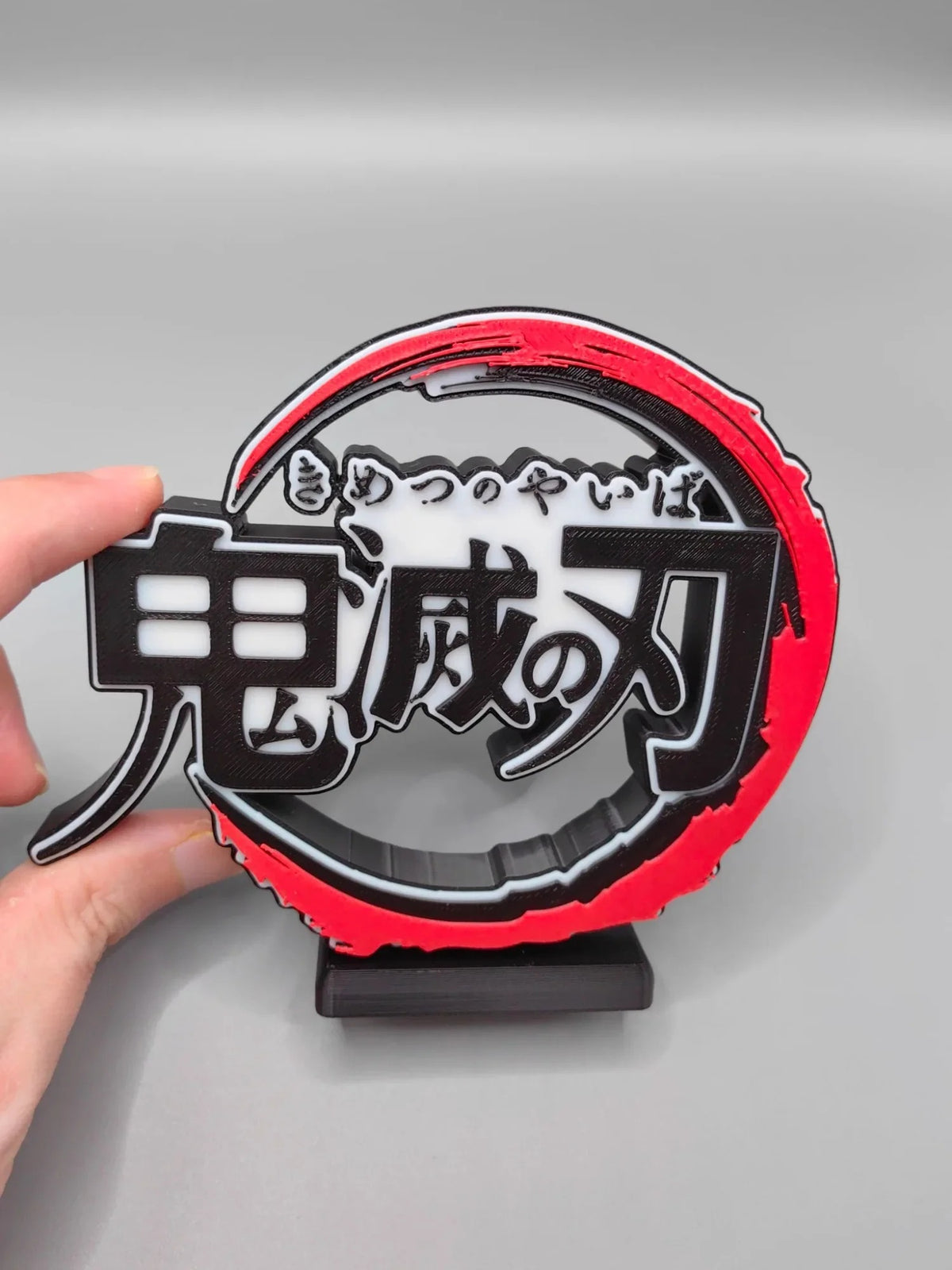 Hand holding a red and black emblem with Japanese text on a gray background Demon Slayer: Kimetsu no Yaiba Premium 3D PVC Logo Model Signboard – 13cm Desktop Title Display Sign for Collectors front