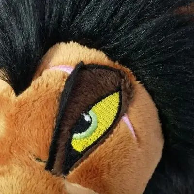 Close-up of a plush toy face showing a large embroidered eye with yellow, green, and black detailing, set against tan plush fabric and black hair. Official The Lion King Scar Premium Cotton Plush by Titantoons face
