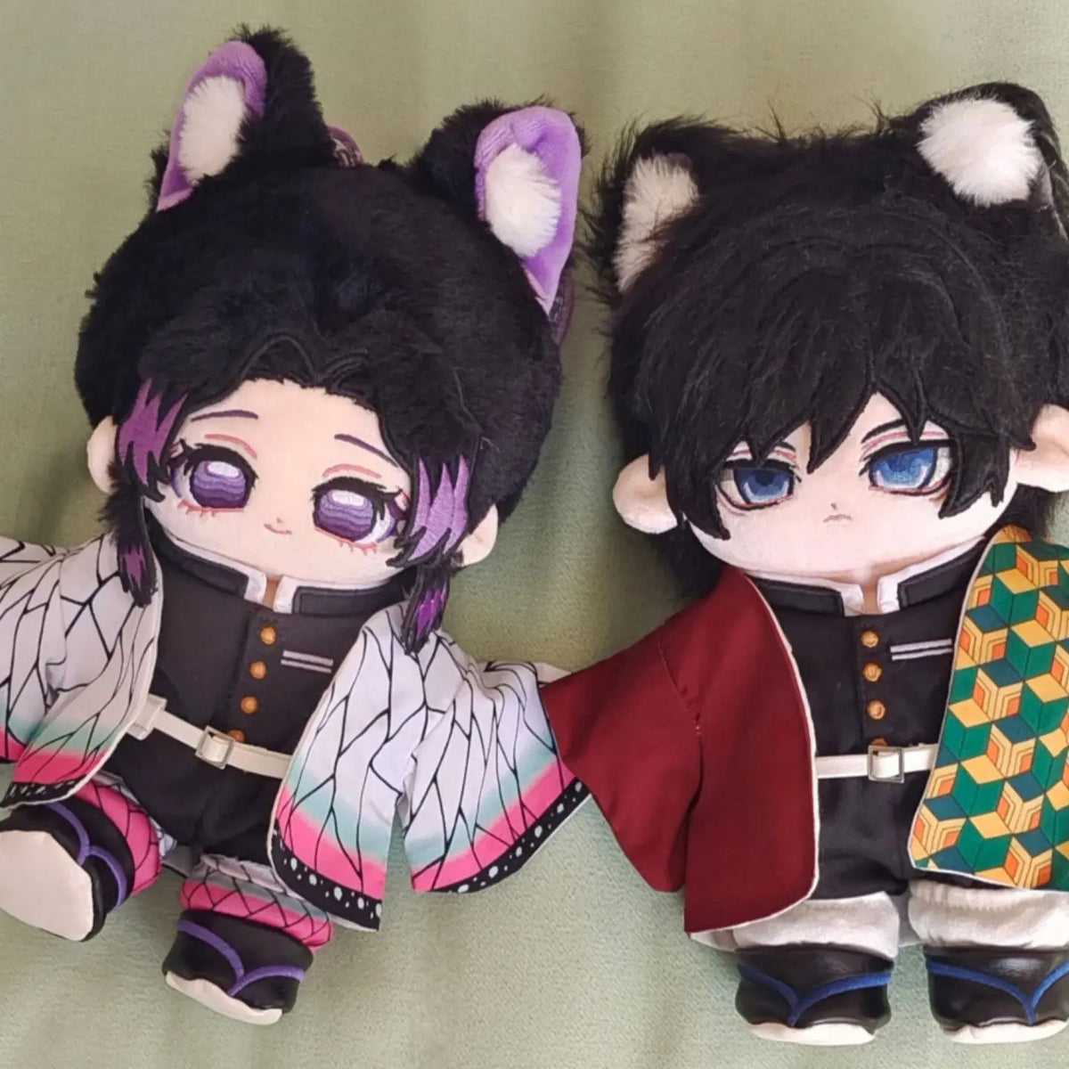 Two plush toys with cat ears, one in black and purple outfit and the other in red and black outfit, on a green background. Demon Slayer: Kimetsu no Yaiba Shinobu Kocho Premium 20cm Super Soft Plushie Doll – Fluffy Purple-Eared Animal Hood Edition duo