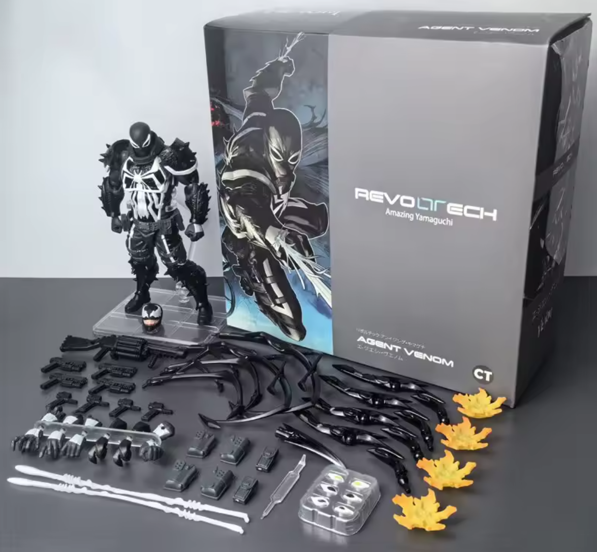 Agent Venom Revoltech action figure set with accessories, shown with its retail box on a gray studio background. The set includes a black-and-white armored figure, multiple black limbs, white weapons, small black accessories, yellow leaf-shaped pieces, and a clear storage tray. Marvel Amazing Yamaguchi Revoltech Agent Venom Authentic Action Figure