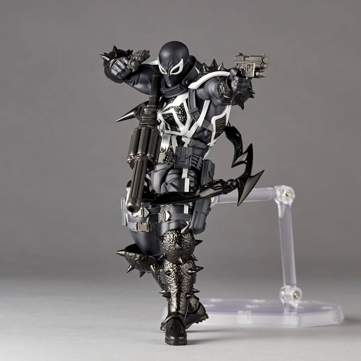 A detailed collectible action figure of Venom in a black and white armored suit, posed dynamically with weapons and accessories, displayed on a clear plastic stand against a neutral gray background. Marvel Amazing Yamaguchi Revoltech Agent Venom Authentic Action Figure