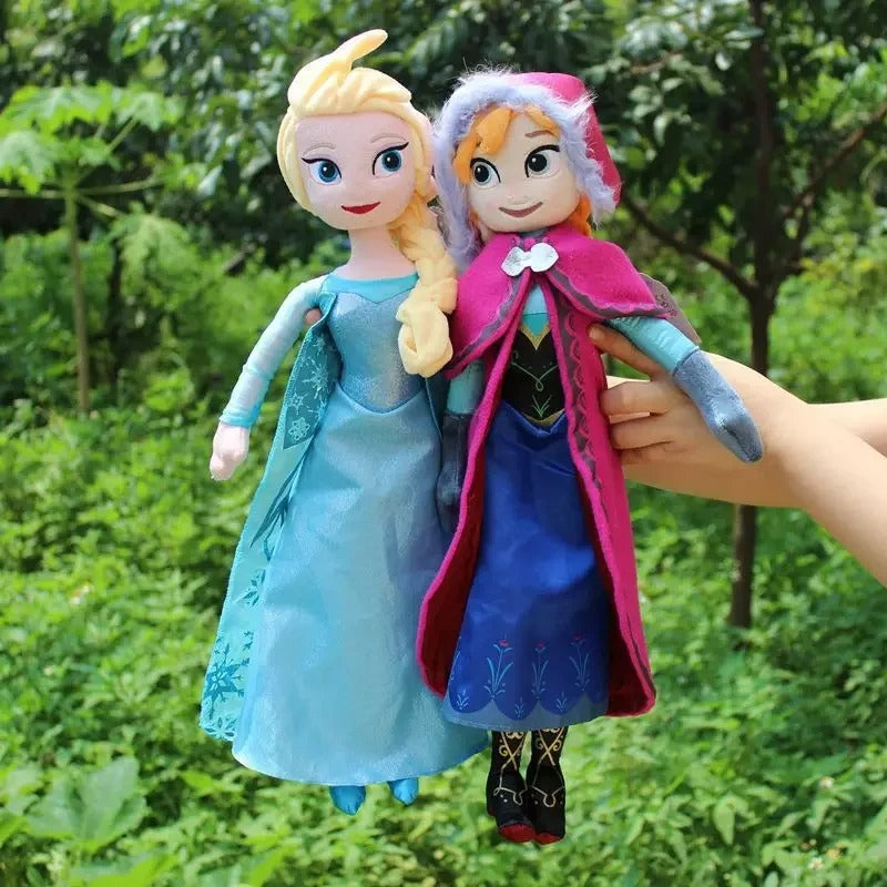 Two dolls, one in a blue dress and the other in a pink cloak, held by a hand against a green outdoor background. Official Disney Frozen Queen Elsa Premium Cotton Plush by Titantoons – Available in 16-Inch & 20-Inch