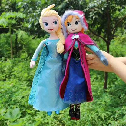 Two dolls, one in a blue dress and the other in a pink cloak, held by a hand against a green outdoor background. Official Disney Frozen Queen Elsa Premium Cotton Plush by Titantoons – Available in 16-Inch & 20-Inch