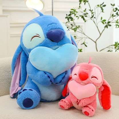 Two plush toys, one blue and one pink, sitting on a light-colored surface with a blurred indoor background. Official Disney Stitch & Angel Hugging Heart Premium Cotton Plush Set by Titantoons