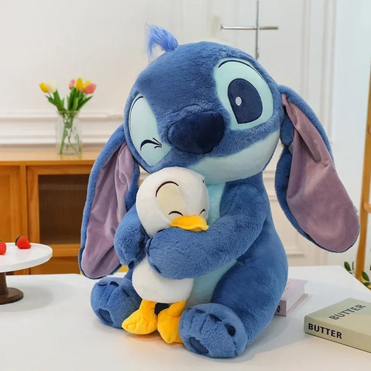 Plush toy of a blue character holding a white duck-like creature on a table with a blurred background. Official Disney Stitch Hugging Duck Premium Cotton Plush by Titantoons – Available in 12-Inch, 18-Inch, & 24-Inch