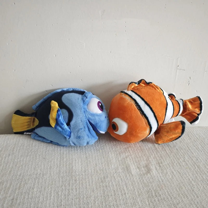 Two plush fish toys from Finding Nemo resting side by side on a textured light beige surface against a plain off-white wall. One plush is blue with yellow and black details, and the other is orange with white stripes and black outlines. Official Finding Nemo & Dory Premium Best Friends Plush by Titantoons