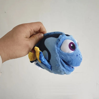 A plush fish toy being held by a hand against a plain light background. The toy is predominantly light blue with dark blue/black patches, a large white eye with a purple iris, and yellow accents. Official Finding Nemo & Dory Premium Best Friends Plush by Titantoons front