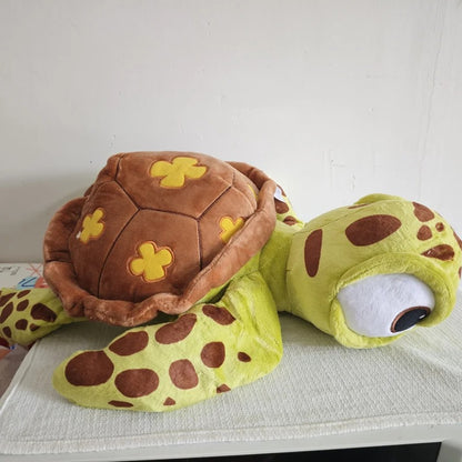 A plush sea turtle toy with a brown shell decorated with yellow flower-like patches and a yellow-green body with brown spots, resting on a white textured surface against a plain light wall. Official Finding Nemo Sea Turtle Premium Cotton Plush by Titantoons 3