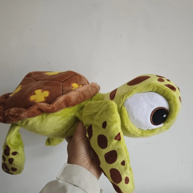 A plush sea turtle toy being held up by a hand against a plain light gray wall. The turtle has a yellow-green body, brown spots, a brown shell with yellow flower details, and a large white eye with a dark pupil. Official Finding Nemo Sea Turtle Premium Cotton Plush by Titantoons