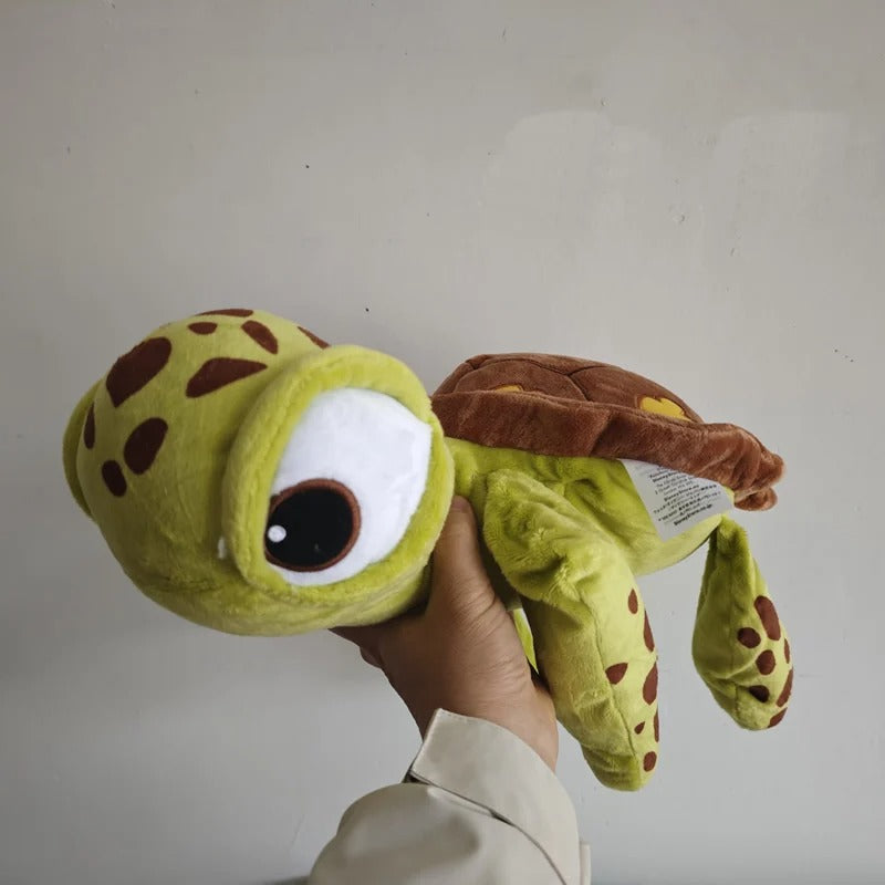 A plush sea turtle toy being held up by a person's hand against a plain light gray wall. The toy is lime green with brown spots, a brown shell, and a large cartoon-style eye. Official Finding Nemo Sea Turtle Premium Cotton Plush by Titantoons side 2