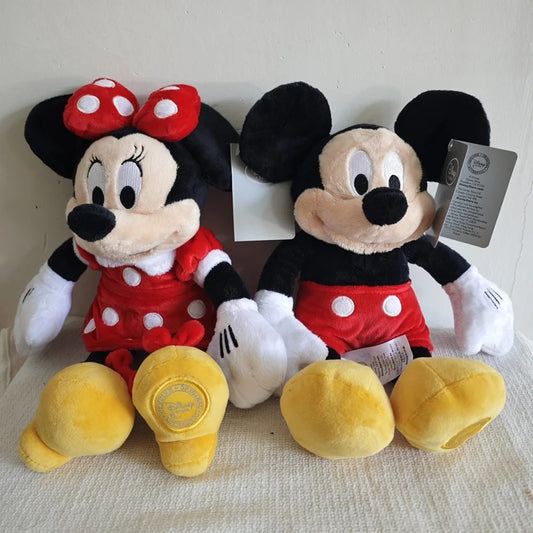 Two plush toys, Minnie Mouse in a red dress and Mickey Mouse in a red shorts set, sitting side-by-side on a textured white surface against a plain beige wall. Official Mickey & Minnie Mouse Premium Cotton Plush Collection by Titantoons