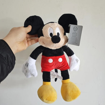 A Mickey Mouse plush toy being held by a hand against a plain off-white wall. The plush has black ears and legs, a beige face, a red shorts with white buttons, white gloves, and yellow shoes. Official Mickey & Minnie Mouse Premium Cotton Plush Collection by Titantoons