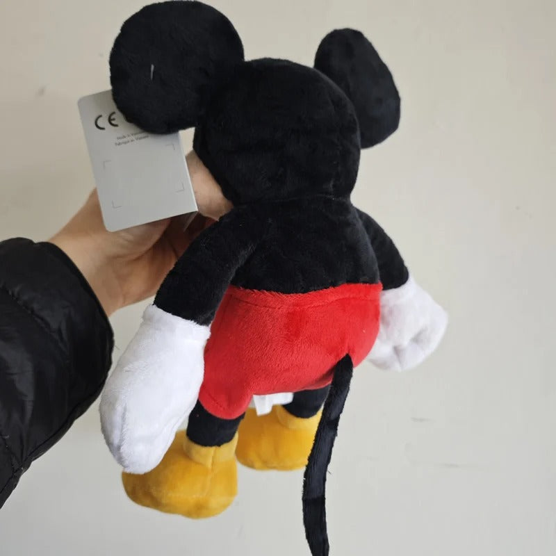 A plush Mickey Mouse toy being held by a hand, showing the back side with its black body, red shorts, white gloves, yellow shoes, and a white hangtag. Official Mickey & Minnie Mouse Premium Cotton Plush Collection by Titantoons
