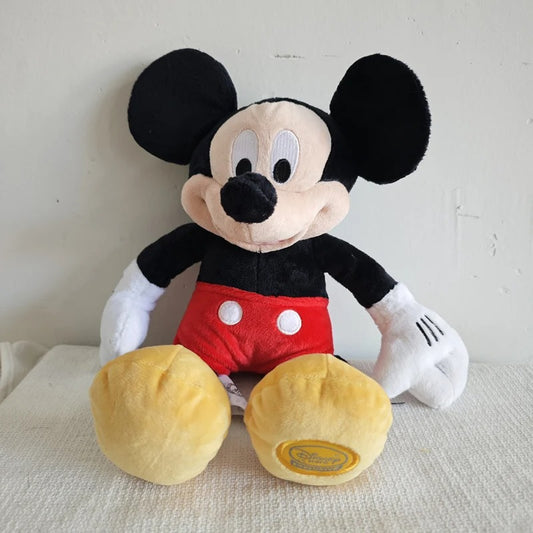 A plush Mickey Mouse stuffed animal sitting on a light fabric surface against a plain off-white wall. The toy features black ears and a black top, red shorts with white buttons, white gloves, and yellow feet. Official Mickey & Minnie Mouse Premium Cotton Plush Collection by Titantoons
