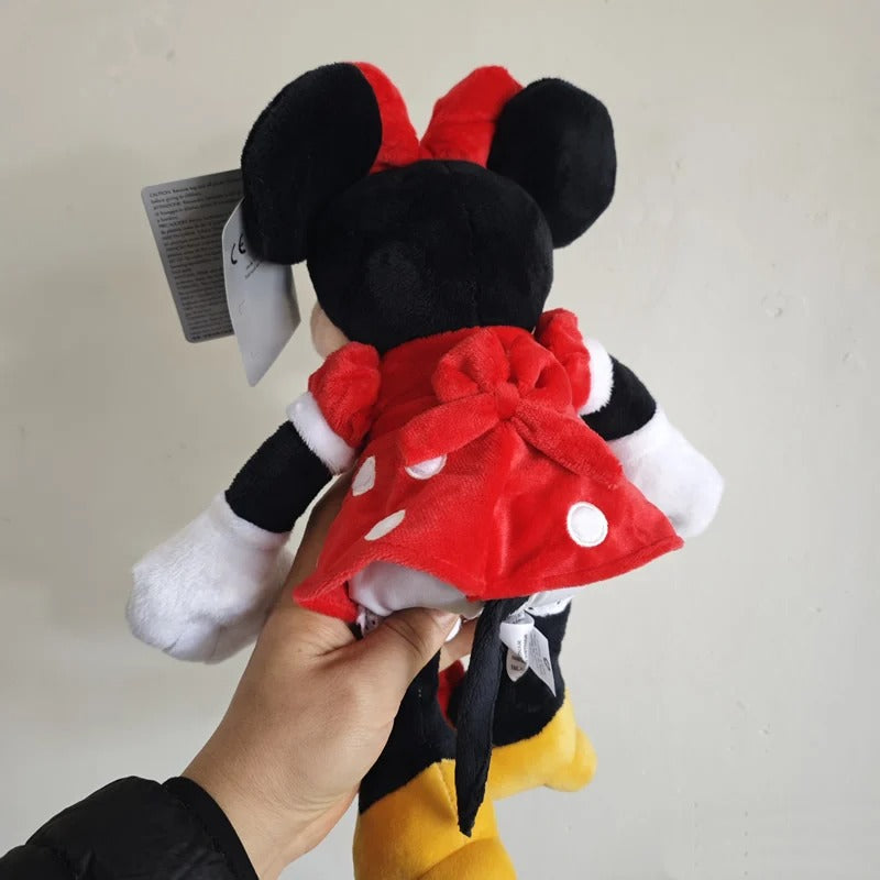 A plush toy of Minnie Mouse being held by a hand against a plain off-white wall. The plush features her signature red polka-dot dress, black ears, white gloves, and yellow shorts, with a grey product tag still attached to her head. Official Mickey & Minnie Mouse Premium Cotton Plush Collection by Titantoons