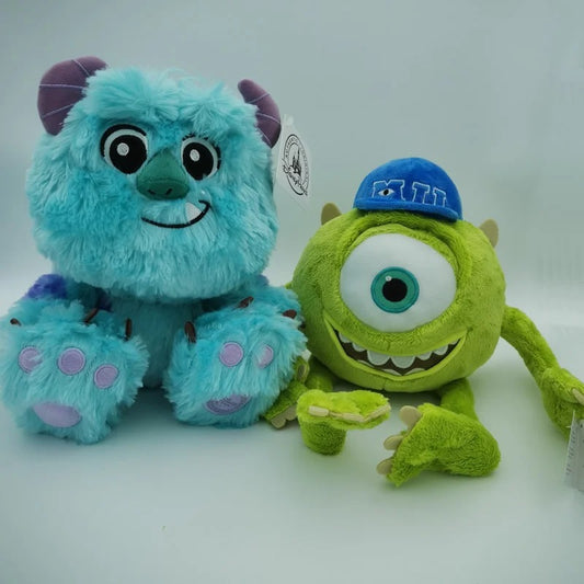 Two plush toys from Monsters Inc. on a white background: a light blue furry Sulley with purple horns and pink paw pads, and a green one-eyed Mike Wazowski wearing a blue cap with 'PUT' embroidered on it. Official Monsters University Sulley & Mike Premium Best Friends Plush Set by Titantoons