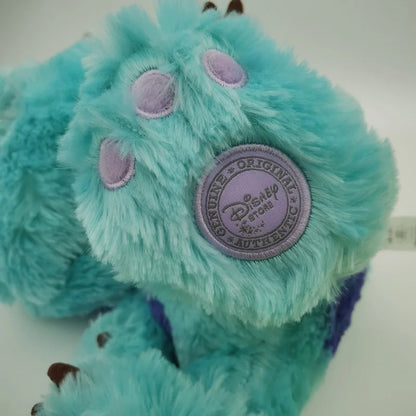 A close-up of a turquoise blue plush monster toy with purple paw pads and a round Disney Collectible Authenticity patch on the side. Official Monsters University Sulley & Mike Premium Best Friends Plush Set by Titantoons bottom