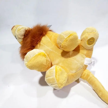 A plush lion toy photographed on a plain white studio background. The toy is mostly pale yellow with a darker brown mane and small white tags attached. Official The Lion King Adult Simba Premium Cotton Plush by Titantoons bottom