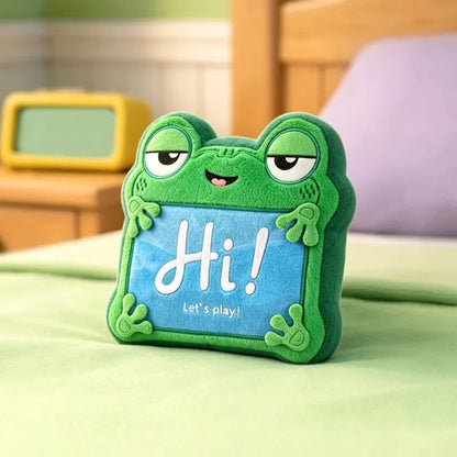 A green plush frog-shaped pillow with a blue front panel that says 'Hi! Let's play!' in white text, photographed on a bed with a wooden headboard and a yellow alarm clock in the background. Official Toy Story & Playroom Friends Premium Cotton Plush Collection by Titantoons