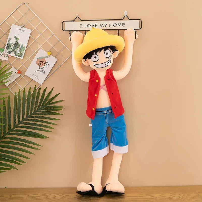 Plush toy of an animated character with a yellow hat and red vest against a beige wall with decorative elements. One Piece: Monkey D. Luffy Giant Luxury Anime Plush