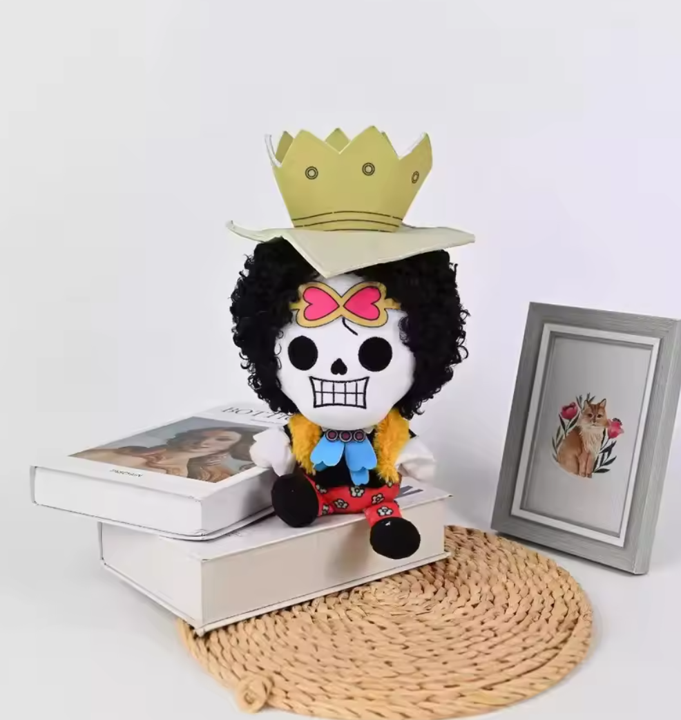 Plush toy with a crown on a white surface with a framed picture and books. One Piece: Soul King Brook Premium 12-Inch Character Plush
